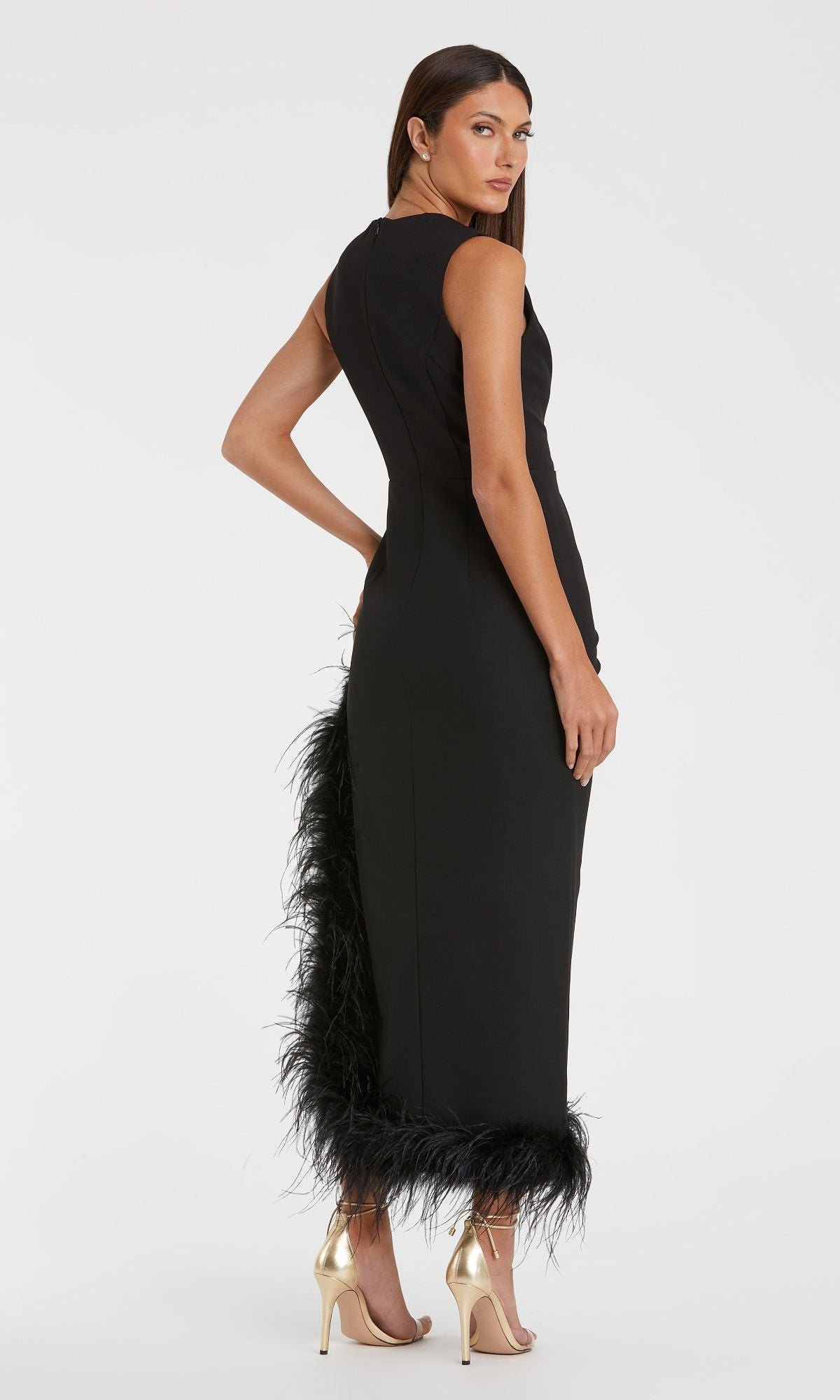 A woman stands sideways, looking back, in the fitted black Mac Duggal 11626 Feather Midi Wedding-Guest Dress with feather-trimmed hem, styled with gold heels and straight dark hair for a striking effect.
